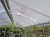 Interior view of a glass conservatory with partially open Exaco Hoklartherm/Exaco Universal Retractable Roll-up Roof Shade Curtains; aluminum-acrylic fabric provides flexible greenhouse shade, with garden plants visible through the glass walls.