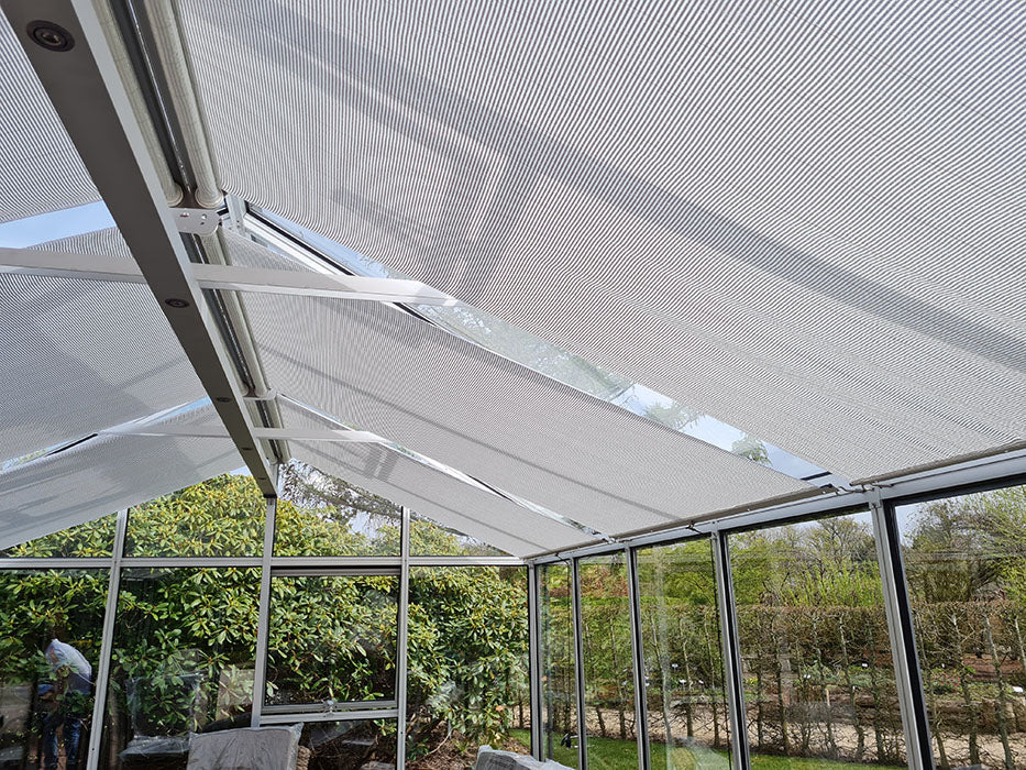 Interior view of a glass conservatory with partially open Exaco Hoklartherm/Exaco Universal Retractable Roll-up Roof Shade Curtains; aluminum-acrylic fabric provides flexible greenhouse shade, with garden plants visible through the glass walls.