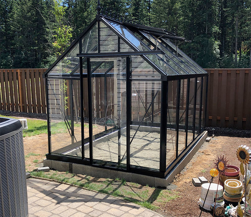 Exaco Janssens Royal Victorian VI 23 greenhouse with black aluminum frame and clear tempered glass panels, set on a foundation in a fenced backyard.