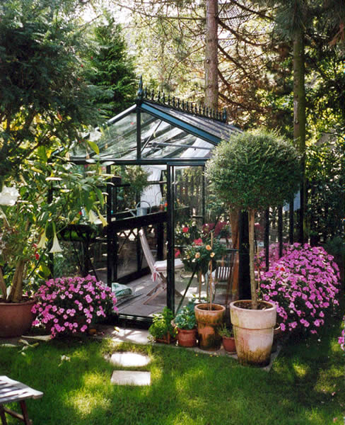 Exaco Janssens Royal Victorian VI23 greenhouse with black frame, surrounded by pink flowers and lush greenery.