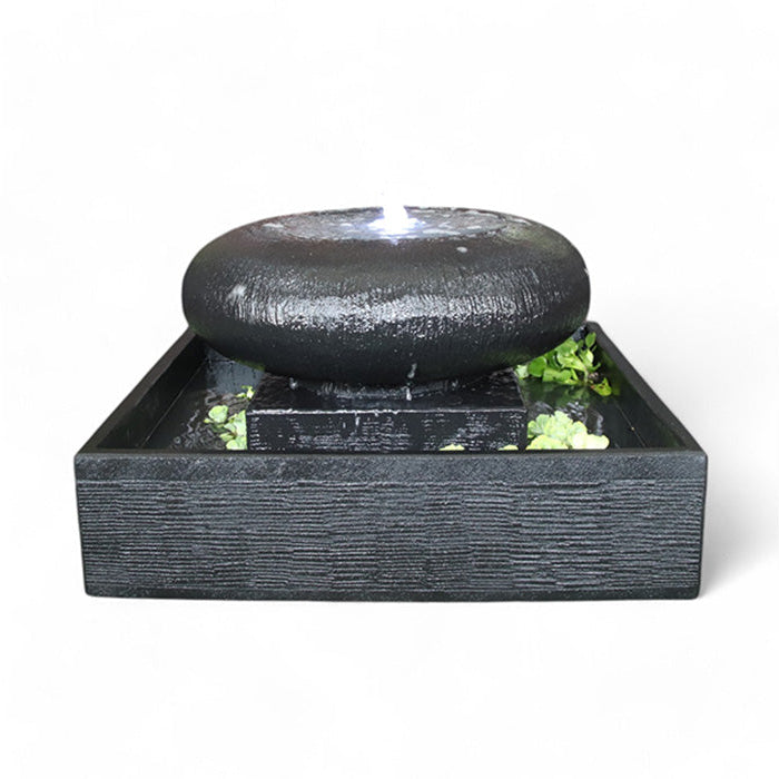 Front view of the Exaco Moon Pot Fountain in a sleek, textured black finish. Water bubbles up from the center of the smooth, round top, illuminated by a white LED light, creating a calming indoor water feature.