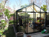 Exaco Royal Victorian VI 34 Greenhouse in a backyard setting with curtain-style shade inside, surrounded by blooming garden plants.