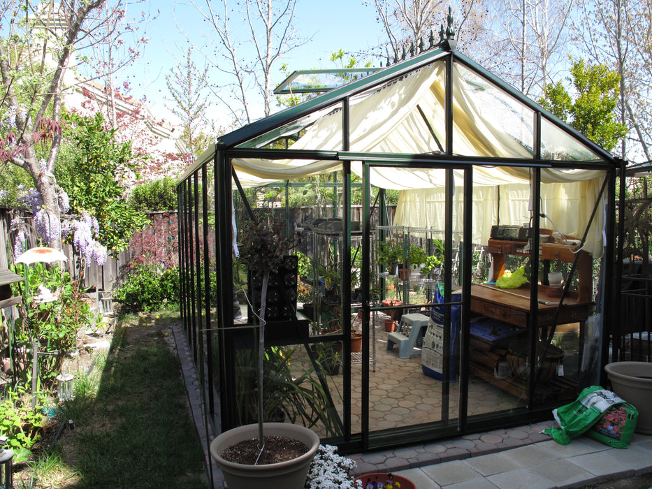 Exaco Royal Victorian VI 34 Greenhouse in a backyard setting with curtain-style shade inside, surrounded by blooming garden plants.