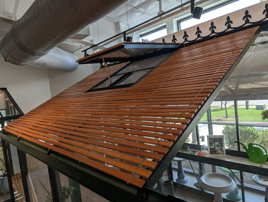 Exaco VI36 greenhouse on showroom floor with wood slatted shades mounted on roof panels, showing interior view with sunlight filtering through.