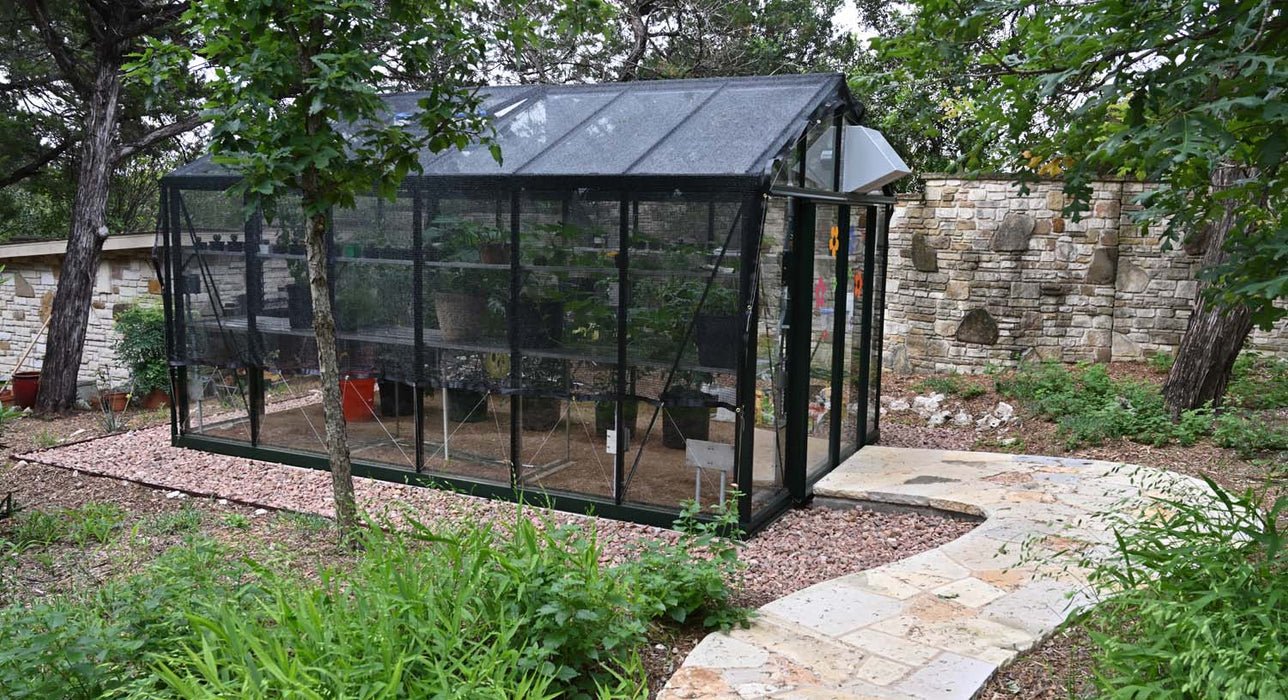 Side view of Exaco Janssens Royal Victorian VI 34 Greenhouse with black shade netting, surrounded by stone path and lush garden plants.