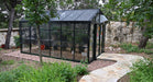 Side view of Exaco Janssens Royal Victorian VI 34 Greenhouse with black shade netting, surrounded by stone path and lush garden plants.