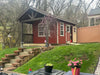 Red 10x16 Ez-Fit Kits Woodsman Shed with 6' covered porch, cupola, open double doors, white-trimmed windows, and landscaped hillside garden