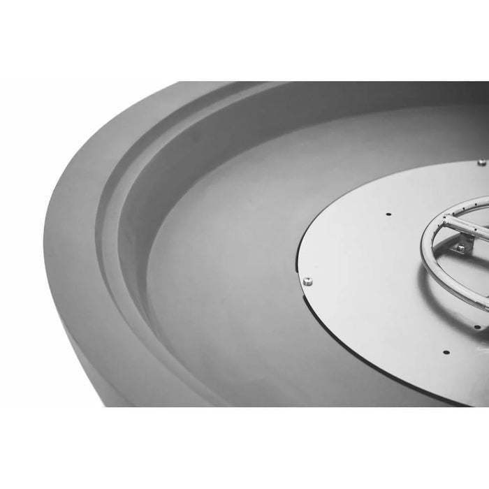Extreme close-up, black-and-white detail shot of a concrete fire bowl's interior rim and round stainless steel burner pan, showing the layered edge profile and drilled ignition ring mounted at the center of the basin.