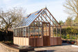 Front angle of a cedar Westminster greenhouse placed in a garden, showing its glass structure, wooden base, and door with a metal handle.