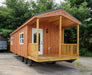 Front view of the Oasis Tiny Estates Firestack Cabin with a spacious porch, wooden railing, and charming entry door, set in a serene natural environment.