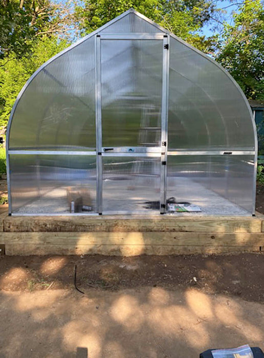 Front entrance view of the Exaco Riga greenhouse with barn-style hinged door, transparent panels, and sturdy aluminum frame on raised wooden base.