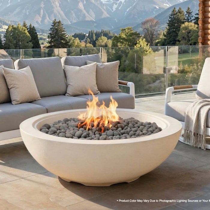 A round frosted cream white concrete fire pit burns brightly with a visible orange flame atop a stone patio, set against a dramatic snow-capped mountain backdrop with a gray sectional sofa, pillows, and a knit throw nearby -- the Elementi Rose Bowl 48 Fire Pit.