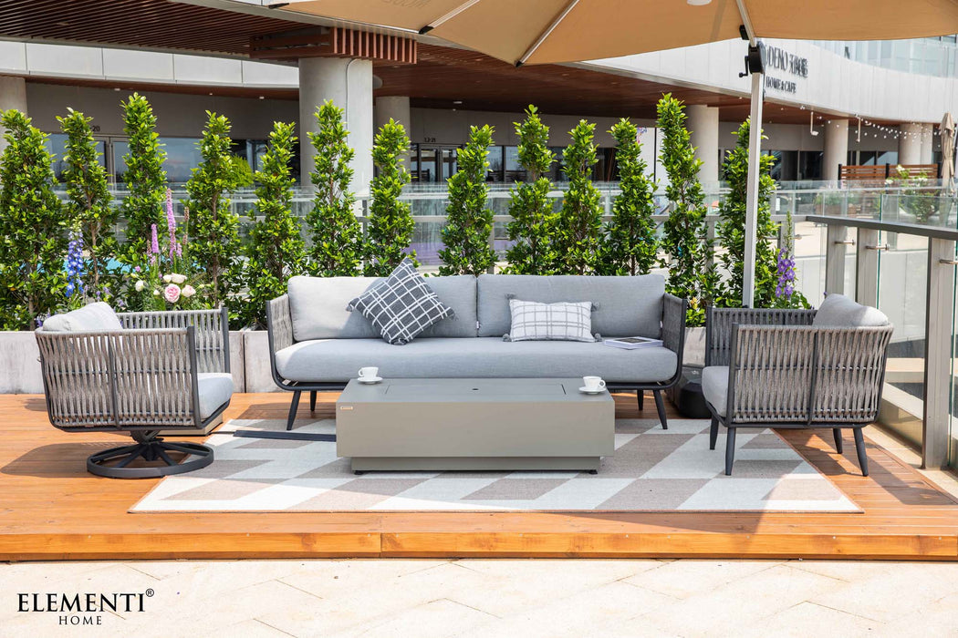 An Elementi Milano Collection outdoor furniture set that includes a grey sofa with patterned pillows, two grey rope swivel armchairs, and a rectangular fire pit table with two coffee cups on top.