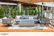 An Elementi Milano Collection outdoor furniture set that includes a grey sofa with patterned pillows, two grey rope swivel armchairs, and a rectangular fire pit table with two coffee cups on top.