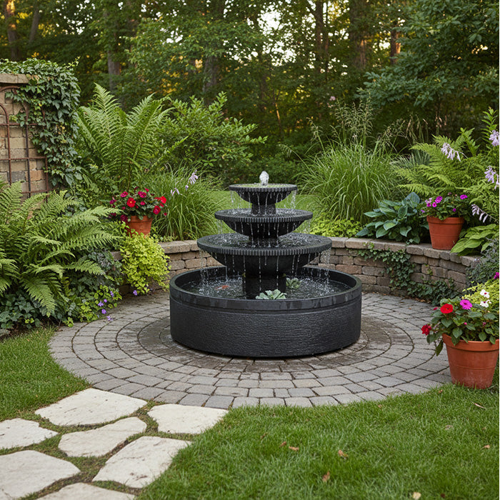 Tiered black outdoor water fountain centered on circular paver patio surrounded by lush green plants and colorful flowers