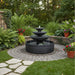 Tiered black outdoor water fountain centered on circular paver patio surrounded by lush green plants and colorful flowers