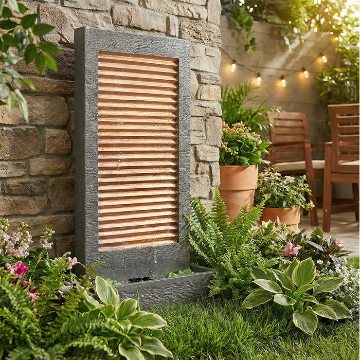 Contemporary garden water feature with copper slat panel and dark textured frame, surrounded by lush plants and grass.
