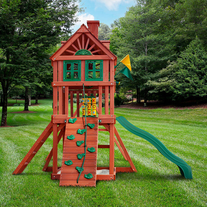 Rear view of the Nantucket II cedar swing set in a lush backyard featuring treehouse roof, green slide, rock wall, sandbox, and tic-tac-toe panel