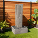 Yang modern water fountain in gray stone with sculpted textured panel and square basin filled with decorative rocks in patio setting.