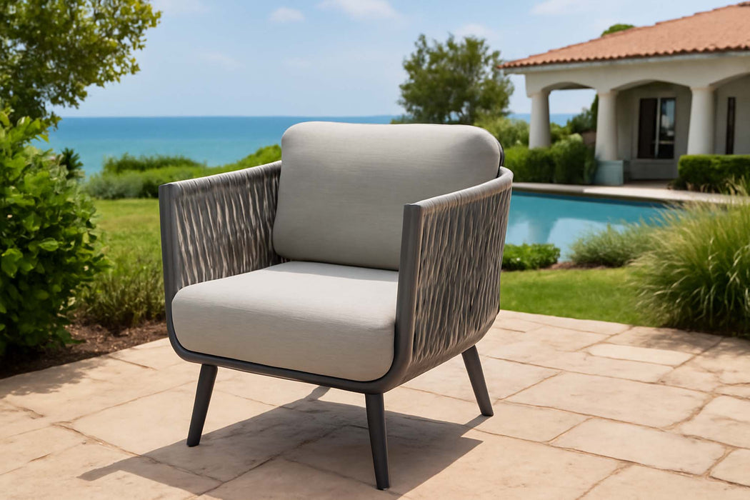 An outdoor armchair with grey rope weave and a grey metal frame, part of the Elementi Milano Collection. It is placed on a stone patio overlooking a swimming pool, a house with a tiled roof, and the ocean in the background under a clear blue sky.