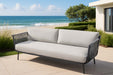 A grey rope weave sofa from the Elementi Milano Collection with two light grey cushions, situated on a stone patio with a direct view of a sandy beach and the ocean with gentle waves.
