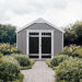 Handy Home 10x8 Acadia with white trim, black double doors, and windows. A stone path leads to the shed through a garden with green shrubs and white flowers.