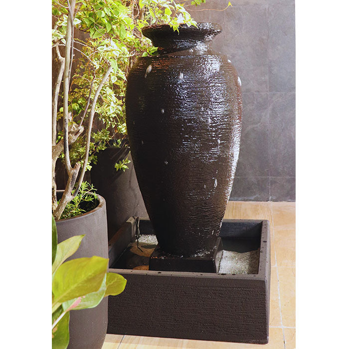 Indoor amphora water fountain with square basin, dark textured finish and warm LED string lights above potted plants.