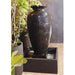 Indoor amphora water fountain with square basin, dark textured finish and warm LED string lights above potted plants.