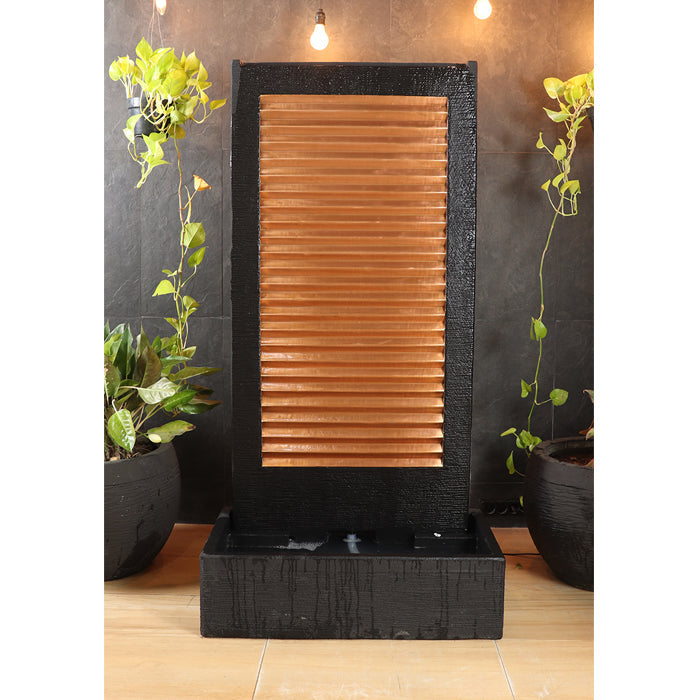 Indoor outdoor black wall fountain with copper slat panel and lower basin, accented by hanging plants and warm lighting.