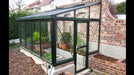 A Janssens Arcadia lean-to greenhouse with a green frame, glass panels, and brick base, attached to a brick wall.