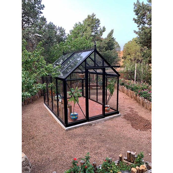 A compact Exaco Janssens Junior Victorian Greenhouse set in a garden, surrounded by rich vegetation and clear skies.