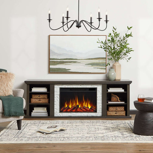 Real Flame Kenai Slim Electric Fireplace TV Stand in a styled living room with landscape painting, black chandelier, armchair, woven baskets, and glowing orange flames