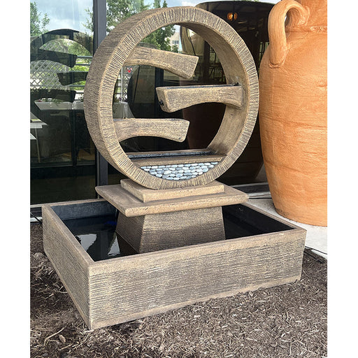 A large, textured, light-colored stone-look Exaco Wagon Wheel Fountain placed outdoors, showcasing its circular tiered water flow design and rectangular base