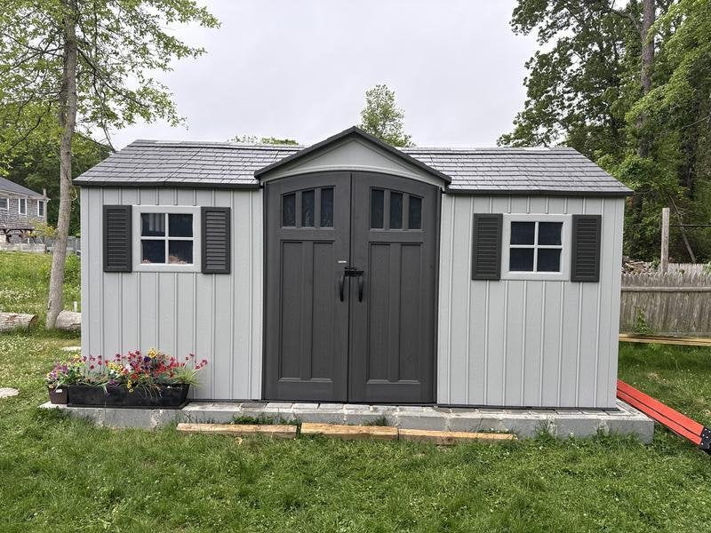 Lifetime 15x8 ft. dual entry storage shed installed in a backyard with flowers, front angle, closed doors.