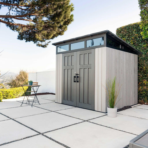 Lifetime Farmhouse Shed 60542 in a modern patio setting with concrete pavers, bistro table, ornamental grass planter, and hillside view under bright sky