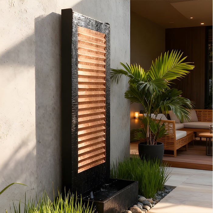 Luxury patio water feature with copper slat panel and black textured frame, surrounded by tropical plants and seating.