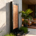 Luxury patio water feature with copper slat panel and black textured frame, surrounded by tropical plants and seating.