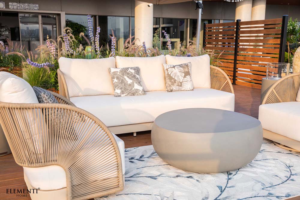 Elementi Outdoor Furnitures Malibu sofa and chair set displayed on a patio with round coffee table, floral cushions, and lush garden surroundings.