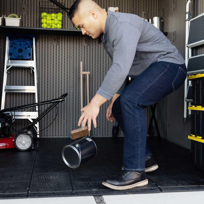 Man bending down to pick up a fallen paint can inside the Lifetime Farmhouse Shed 60542, showing impact-resistant dark floor, lawn mower, ladder, and wall shelving