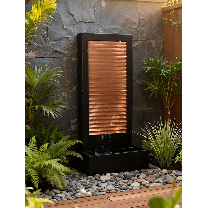 Modern black and copper outdoor wall fountain with rectangular basin, placed among river rocks and garden plants.
