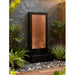 Modern black and copper outdoor wall fountain with rectangular basin, placed among river rocks and garden plants.