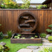 Modern circular cascading outdoor water fountain in brown resin finish with tiered spouts and square basin, styled in a serene backyard garden setting.
