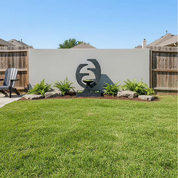 Wide backyard view highlighting Eclipse modern outdoor fountain against stucco wall with green lawn and decorative rocks.
