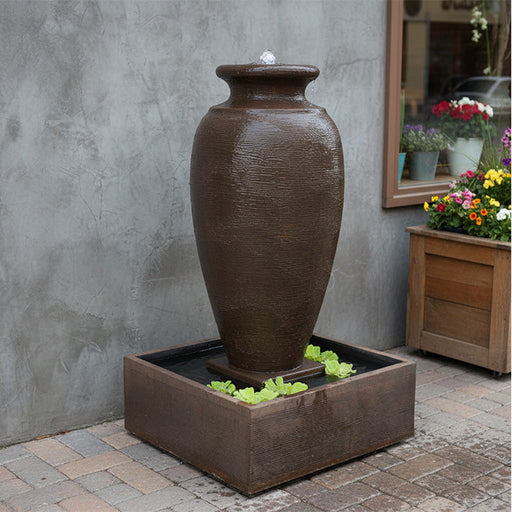 Modern garden amphora fountain with square water reservoir, bubbling top and lush greenery surrounding backyard lawn.