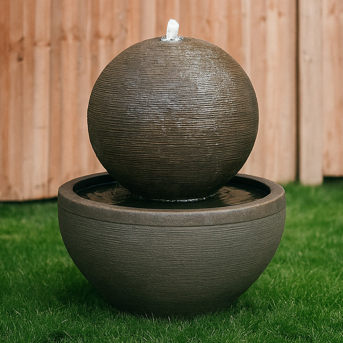 Modern Luna Bowl water fountain with ribbed spherical top and deep round basin, displayed on grass with wooden backdrop.