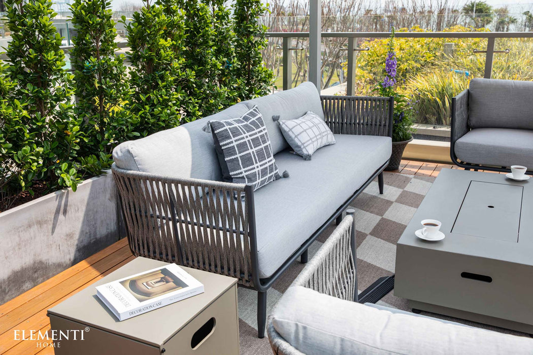 A modern Elementi Milano Collection outdoor seating area featuring a light grey sofa with a dark grey checkered and a light patterned accent pillow, along with a glimpse of matching armchairs. 