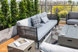 A modern Elementi Milano Collection outdoor seating area featuring a light grey sofa with a dark grey checkered and a light patterned accent pillow, along with a glimpse of matching armchairs. 