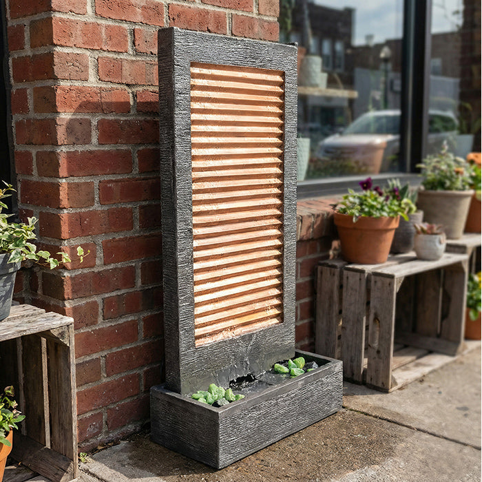 Modern outdoor wall fountain with horizontal copper panel and dark frame, placed against brick wall with planter base.