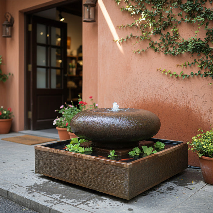 Moon bowl water fountain with square base placed on patio near stucco wall and potted flowers, creating a modern outdoor accent.