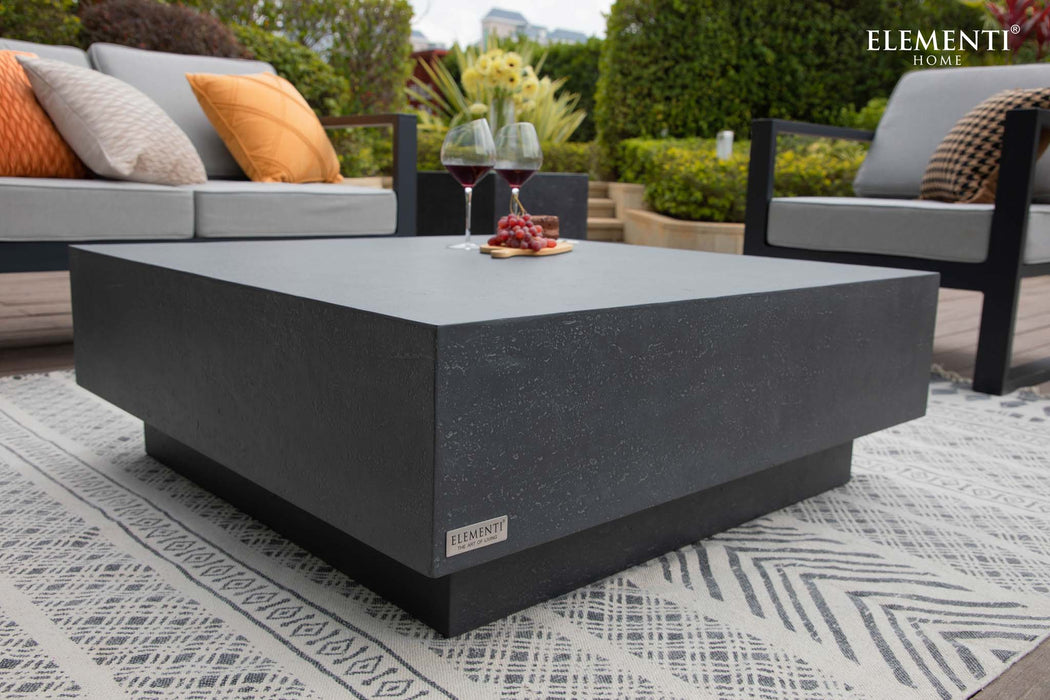 A stylish outdoor seating arrangement featuring the Elementi Tevere Series Square Coffee Table in Slate Black, with wine and snacks on the table.